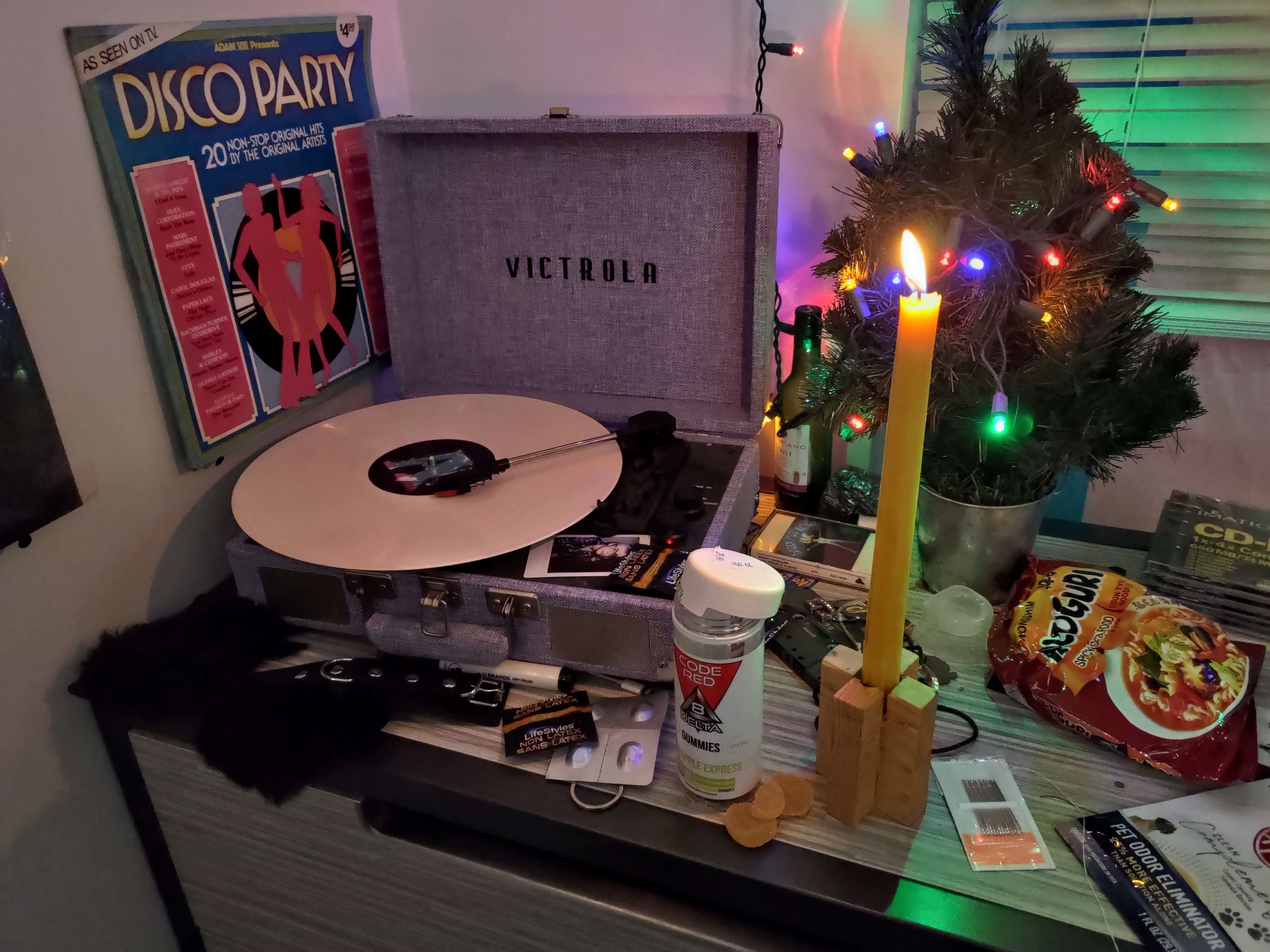 An image of a record player on a cluttered surface. It has multiple psychoactive substances and sex toys visable alongside it, as well as a small plastic x-mas tree.