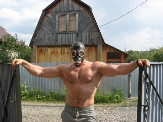 unbelievably buff man in a gas mask standing at the gate of a farm with a barn